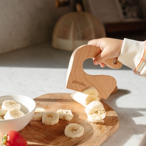 May include: A child's hand holds a wooden banana slicer with the text "piccalio." on it. The slicer is being used to cut a banana on a wooden cutting board. There are banana slices on the board and in a white bowl. There is a strawberry on the board.