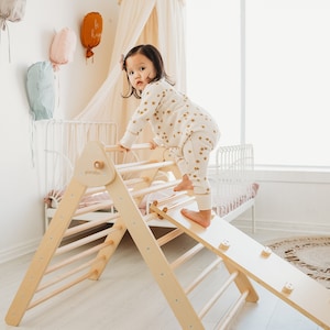 May include: A wooden toddler climbing triangle with a slide. The triangle has a ladder on one side and a slide on the other. A young child is climbing up the ladder. The triangle is made of natural wood and has a light brown finish. The slide is made of a smooth, light-colored wood. The child is wearing a white and gold star patterned shirt and pants.