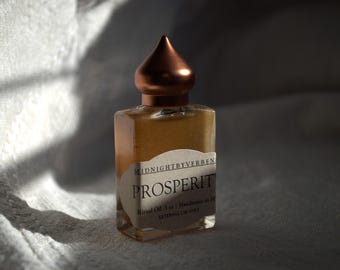 May include: A small amber glass bottle with a copper-colored cap and a label that reads "MIDNIGHTBYVERBEN PROSPERIT Ritual Oil 3 ml | Handmade in LA External Use Only 2017"
