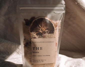 May include: A clear plastic bag filled with a beige bath salt blend. The bag has a label that reads "MIDNIGHTBYVERBENA THE lovers passion | romance | love drawing Botanical Salt Bath 7 oz Handcrafted in MS". A dried orange slice is visible in the bath salt.