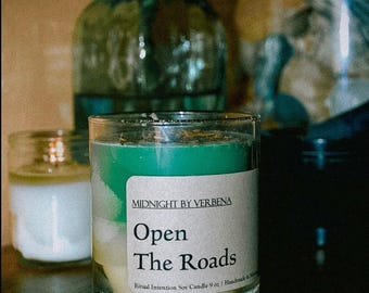 May include: A green glass candle jar with a white label that reads "Midnight by Verbena Open The Roads Ritual Intention Soy Candle 9 oz | Handmade in Phoenix".