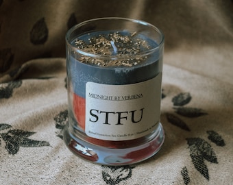 May include: A black and red soy candle with the text "STFU" and "Midnight by Verbena" on the label. The candle is in a clear glass jar and has a gold glitter top.