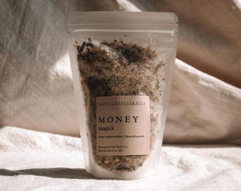 May include: A clear plastic pouch filled with a white and brown bath salt blend. The label reads "MONEY magick attract opportunities | financial growth Botanical Salt Bath Tea Handcrafted in MD"