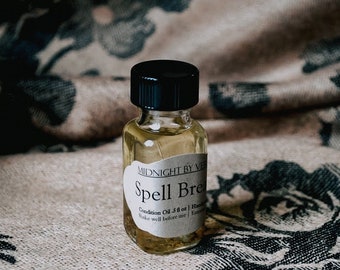 May include: A small, clear glass bottle with a black lid, containing a yellow liquid. The bottle has a white label that reads "Midnight by Vex" and "Spell Brew Condition Oil 5 fl oz | Handcrafted | Essential Oils | Shake well before use".
