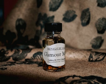 May include: A small glass bottle with a black cap filled with a golden liquid. The label reads "MIDNIGHT BY VER Glamour M Condition Oil 5 fl oz | Handmade | Shake well before use | External"