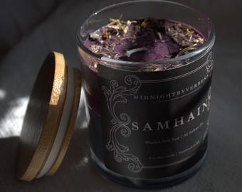 May include: A purple candle with a black label that reads "MIDNIGHT BY VERBENNA SAMHAIN Witches New Year | All Hallows Eve 9 oz Soy Candle | Handmade in the USA"
