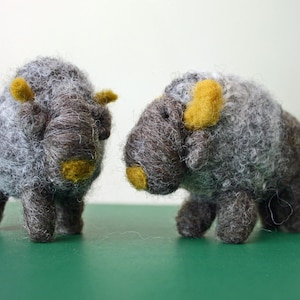 May include: Two handmade felt bison figurines. The bison are gray with brown details and yellow accents on their horns and noses. They stand on four legs and have small tails. The background is a solid green.