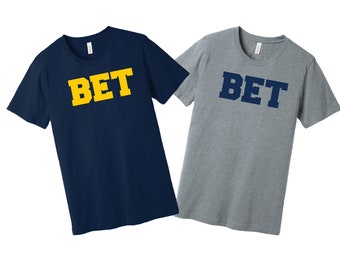 May include: Two t-shirts with the word "BET" printed on the front. One shirt is navy blue with yellow lettering, and the other is gray with navy blue lettering.