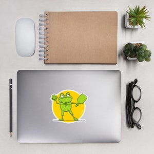 Puede incluir: Un sticker con una rana verde de dibujos animados que sostiene una pelota amarilla y una pala verde, de pie sobre un círculo amarillo. La rana lleva una camisa y pantalones cortos verdes.