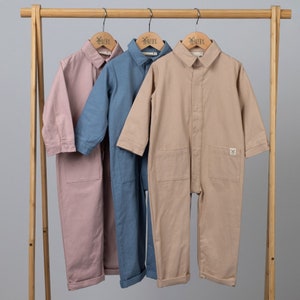 Aneby boilersuits in pink, blue and beige, hanging from wooden clothes frame