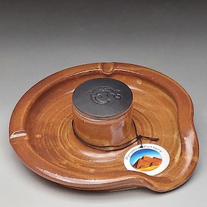 Könnte beinhalten: Ein brauner Keramik-Aschenbecher mit einem zentralen zylindrischen Behälter und einem Deckel. Der Aschenbecher hat ein kreisförmiges Design mit einer gebogenen Kante und einer kleinen Kerbe zum Ablegen von Zigaretten. Ein Etikett mit dem Text "Pyramid Pottery" ist angebracht.