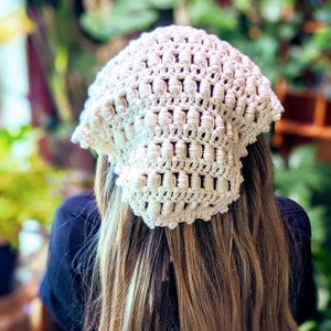 May include: A cream-coloured, handmade crochet hat with a textured, open weave design. The hat is square-shaped and appears to be made of soft yarn. The hat is worn on a person's head, with long, light brown hair visible.