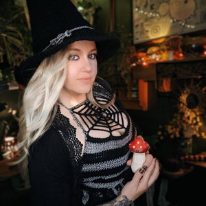 May include: A black crochet witch hat with a silver band and a matching crochet top with a spiderweb design. The person is holding a red and white mushroom figurine. The outfit is a Halloween costume.