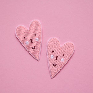 Peut inclure: Deux cœurs en feutre rose avec des visages souriants brodés sur un fond rose.