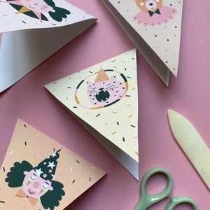 May include: A set of five triangle-shaped paper party banners with a pink, beige, and black confetti pattern. Each banner features a different cartoon animal wearing a party hat: a girl, a bear, a cat, and two clowns. The banners are on a pink surface with a pair of scissors and a craft knife.