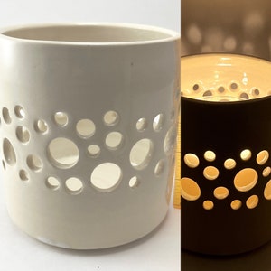 May include: A white ceramic candle holder with a pattern of circles cut out. The candle holder is lit and the light shines through the holes.