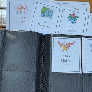 May include: A black card binder with several trading cards featuring colorful illustrations of various creatures. The cards are labeled with numbers and names, including "Goldeen," "Bulbasaur," "Ivysaur," and "Moltres."