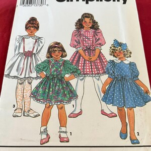 Könnte beinhalten: Simplicity Schnittmuster Nummer 3273 für Mädchenkleider. Das Schnittmuster enthält vier verschiedene Kleidstyles: ein weißes Kleid mit Peter-Pan-Kragen und Puffärmeln, ein grünes Blumenkleid mit Peter-Pan-Kragen und Puffärmeln, ein pink-weißes Karo-Kleid mit Peter-Pan-Kragen und Puffärmeln und ein blaues Punktekleid mit Peter-Pan-Kragen und Puffärmeln.