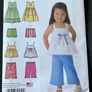 May include: Simplicity easy-to-sew pattern for girls' clothing. The pattern includes options for dresses, shorts, and pants. The image shows a girl wearing a white eyelet top with a blue bow and blue pants. The pattern is designed and printed in the USA.