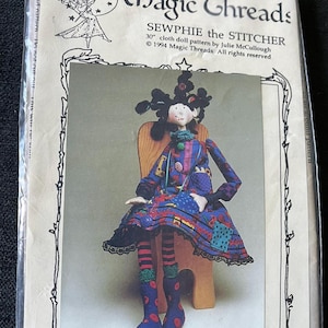 May include: A package for a cloth doll pattern titled "Magic Threads SEWPHIE the STITCHER." The pattern includes a 30-inch doll design. The doll in the image is seated on a wooden chair, wearing a patchwork dress and boots.