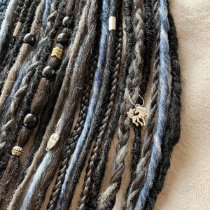 Pode incluir: Um close-up de um conjunto de dreadlocks sintéticas pretas e azuis com contas e pingentes prateados. As dreadlocks estão dispostas em uma fila vertical e têm uma variedade de texturas.