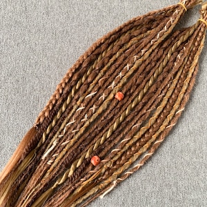 Könnte beinhalten: Braune und blonde synthetische Dreadlocks mit goldenen und weißen Akzenten. Die Dreadlocks haben zwei rote Perlen.