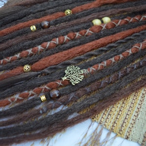 May include: Close-up of brown and reddish-brown synthetic dreadlocks with gold and brown beads and a tree-shaped charm. The dreadlocks are arranged in rows and are tied with brown string.
