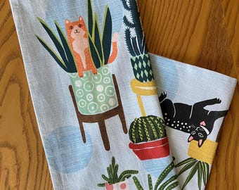 May include: Two light blue kitchen towels with a cat and plant design. One towel features a ginger cat in a green pot, and the other has a black cat. The towels have a whimsical illustration of plants and cats.