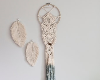 May include: A macrame wall hanging with a 7.5 inch ring and a length of 34 inches. The hanging is made with natural colored cord and has a blue ombre effect at the bottom. The hanging is displayed on a white wall with two smaller macrame feathers.