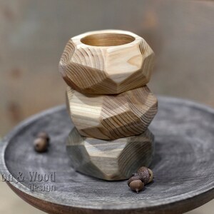 May include: Three wooden geometric candle holders stacked on top of each other. The holders are made of natural wood with a light brown color. The holders are on a dark gray wooden surface.