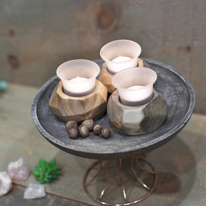 May include: Three small glass candle holders with white candles inside, sitting on a dark gray wooden tray with a wire stand. The tray is decorated with small brown acorns.