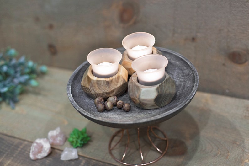 May include: Three small glass candle holders with white candles inside, sitting on a dark gray wooden tray with a wire stand. The tray is decorated with small brown acorns.