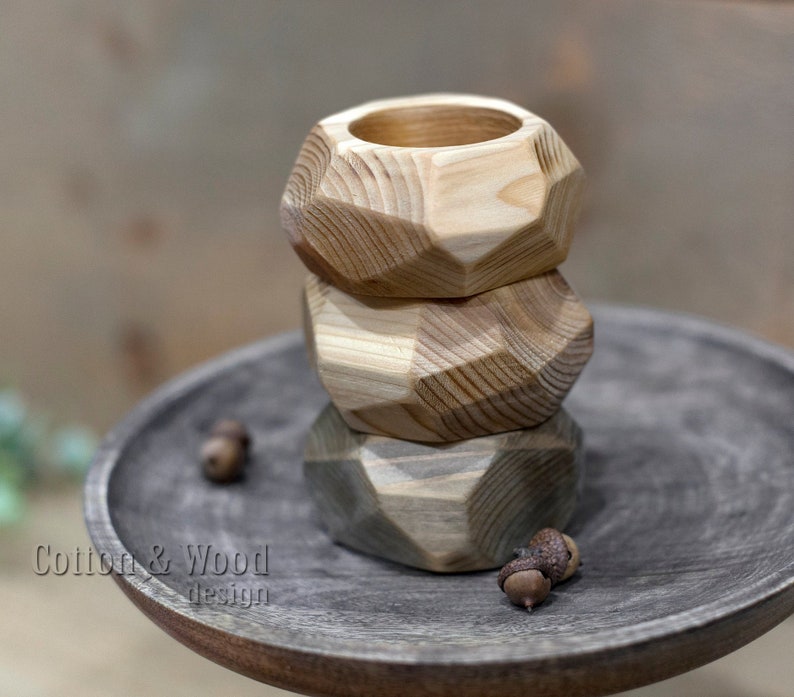 May include: Three wooden geometric candle holders stacked on top of each other. The holders are made of natural wood with a light brown color. The holders are on a dark gray wooden surface.