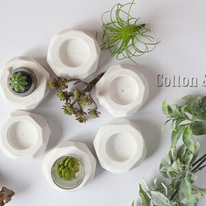 May include: Six white geometric concrete candle holders with a small succulent plant in one of the holders. The holders are arranged in a circle on a white surface. The text "Cotton & Wood design" is visible in the upper right corner of the image.