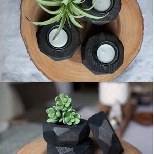 May include: Three black wooden candle holders with a geometric design. The candle holders are on a wooden slice. The candle holders are filled with white tea lights. A small green succulent plant is in the center of the wooden slice.