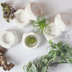 May include: White geometric candle holders with a textured finish. The holders are arranged on a white surface with green plants.