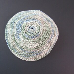 May include: A handmade, crocheted kippah in a circular shape. The kippah features a spiral design with a blend of colors, including white, blue, and green. The kippah is made of yarn and is suitable for religious or cultural purposes.
