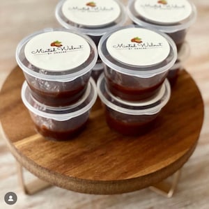 May include: A collection of small, clear plastic containers filled with a dark brown substance. Each container has a white circular lid featuring a logo with a walnut illustration and the text Minted Walnut BY DENISE printed in black.