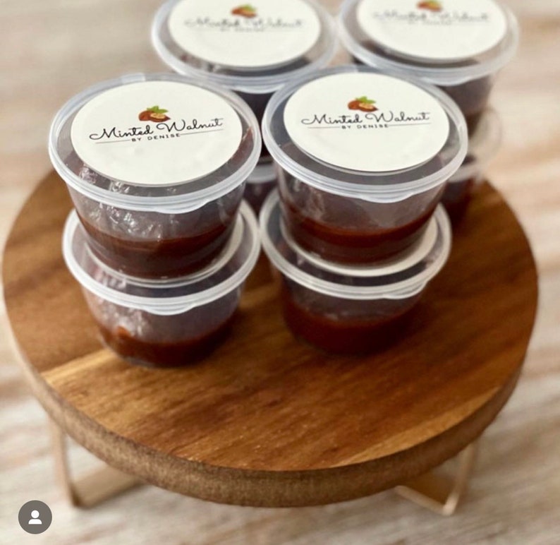 May include: A collection of small, clear plastic containers filled with a dark brown substance. Each container has a white circular lid featuring a logo with a walnut illustration and the text Minted Walnut BY DENISE printed in black.