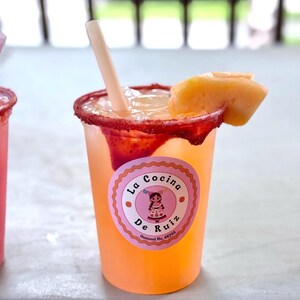 May include: A clear plastic cup filled with a light orange beverage, ice, and a straw. The rim is coated in red chili powder. A slice of cantaloupe sits on the edge. A round pink sticker features the text La Cocina De Ruiz Osmond Ne, 68765.