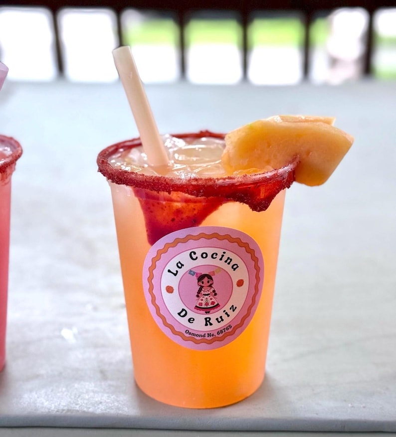 May include: A clear plastic cup filled with a light orange beverage, ice, and a straw. The rim is coated in red chili powder. A slice of cantaloupe sits on the edge. A round pink sticker features the text La Cocina De Ruiz Osmond Ne, 68765.