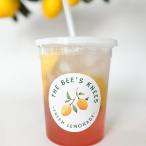 May include: A clear plastic cup filled with pink lemonade, ice cubes, and lemon slices, topped with a white lid and straw. A circular label on the front features two yellow lemons on a branch and the text THE BEE'S KNEES FRESH LEMONADE.
