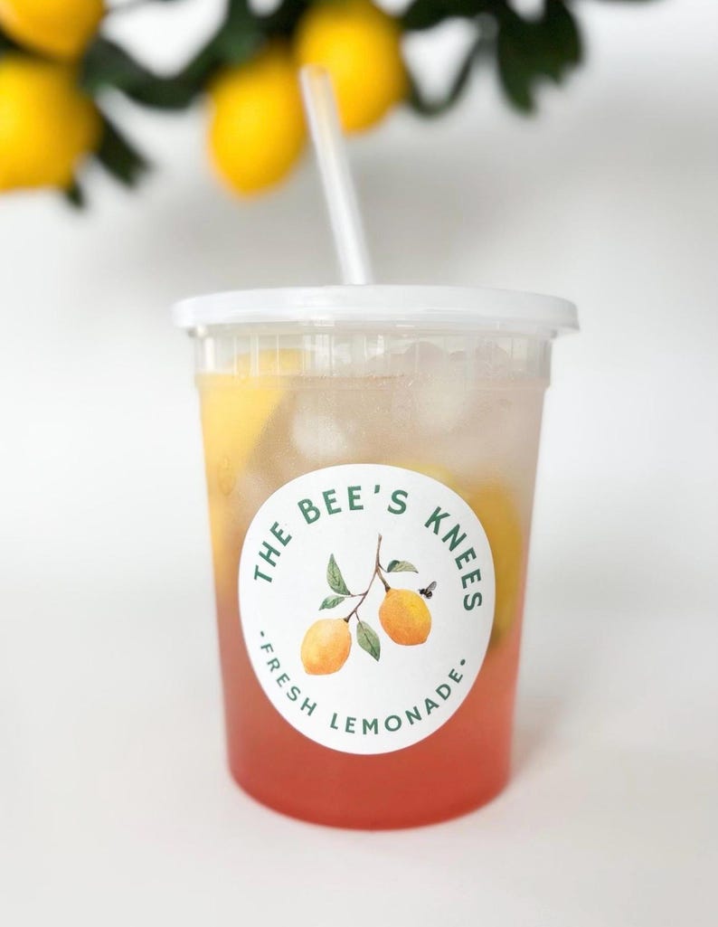 May include: A clear plastic cup filled with pink lemonade, ice cubes, and lemon slices, topped with a white lid and straw. A circular label on the front features two yellow lemons on a branch and the text THE BEE'S KNEES FRESH LEMONADE.
