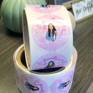 May include: A roll of circular adhesive stickers featuring a pink background with a repeating illustration of a person with long dark hair wearing a blue jacket and holding a pink cup. The text Beecraftyxo is printed above the illustration.