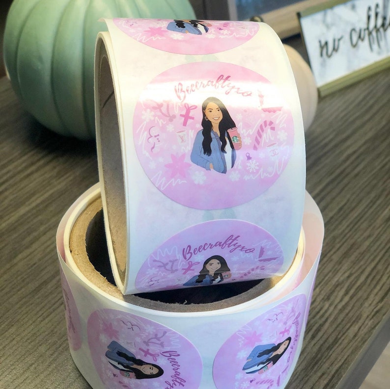 May include: A roll of circular adhesive stickers featuring a pink background with a repeating illustration of a person with long dark hair wearing a blue jacket and holding a pink cup. The text Beecraftyxo is printed above the illustration.