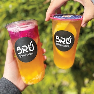 May include: Two clear plastic cups filled with fruit tea, each featuring a black circular sticker with the white text BRU and the Instagram handle BRUTEACAFE. The left cup contains pink dragon fruit and yellow fruit pieces, while the right cup holds orange slices.
