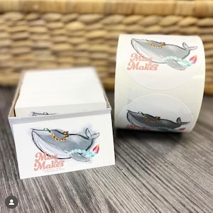 May include: A stack of rectangular white business cards and a roll of circular stickers. Both feature a gray whale illustration wearing a colorful floral lei and light blue beads, with the pink text Miss Lou Makes printed on the front.