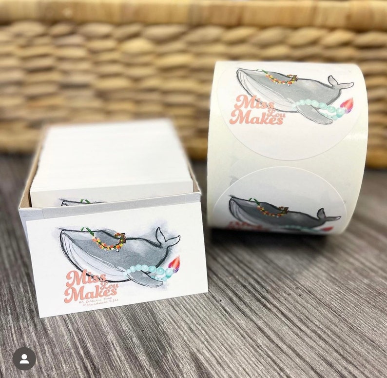 May include: A stack of rectangular white business cards and a roll of circular stickers. Both feature a gray whale illustration wearing a colorful floral lei and light blue beads, with the pink text Miss Lou Makes printed on the front.