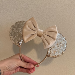 May include: A pair of gold sequin mouse ears with a large beige velvet bow. The ears are attached to a brown headband.