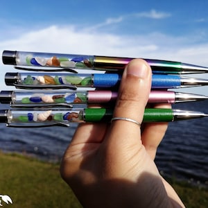 Peut inclure: Cinq stylos avec des barils transparents remplis de verre de mer coloré et de coquillages. Les stylos ont des capuchons métalliques de différentes couleurs : arc-en-ciel, bleu, rose et vert.